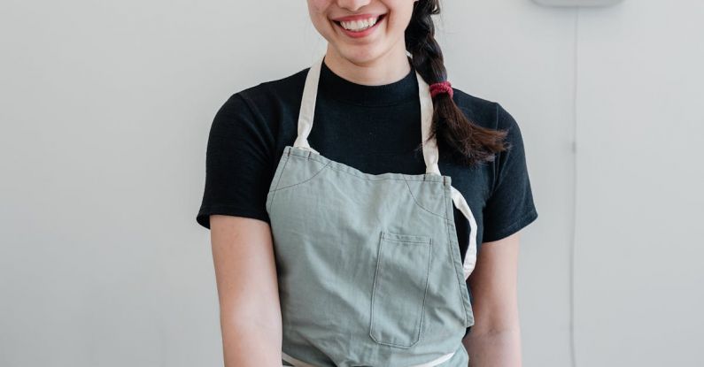 Local Café - Positive young Asian woman wearing apron giving paper cup with coffee to go while standing at wooden counter during work in modern coffee house and looking at camera with smile
